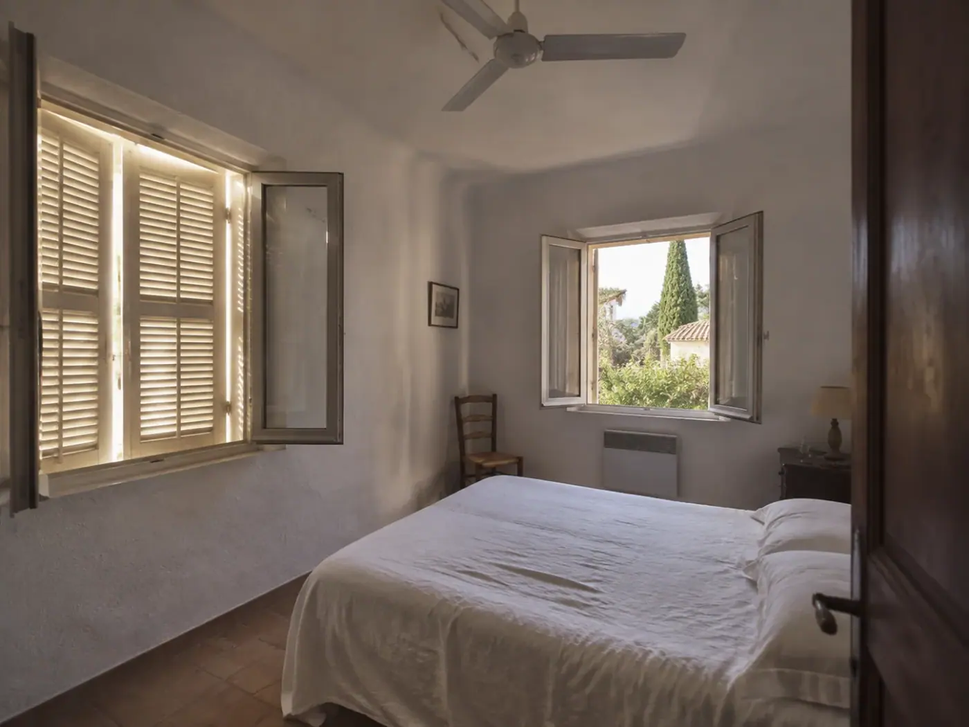 Chambre avec deux fenêtres sur des murs différents, des persiennes partiellement fermées et un ventilateur de plafond.