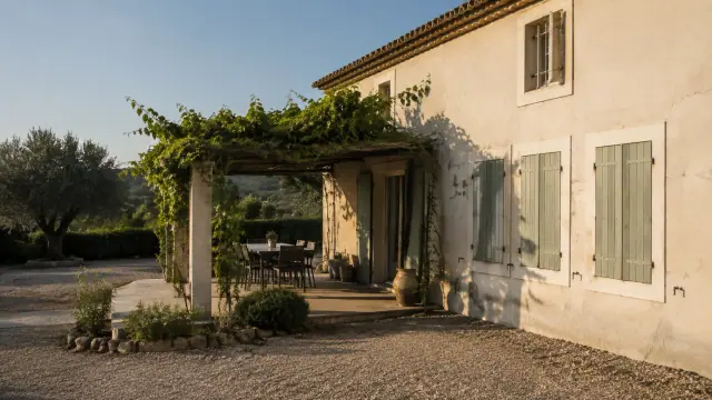 Façade d’une maison aux volets fermés, terrasse ombragée par une pergola végétalisée et jardin de gravier.